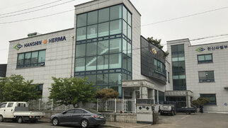 Moderne Bürogebäude mit großen Fenstern, beschriftet mit "HANSHIN" und "HERMA". Umgeben von Bäumen und parkenden Fahrzeugen.