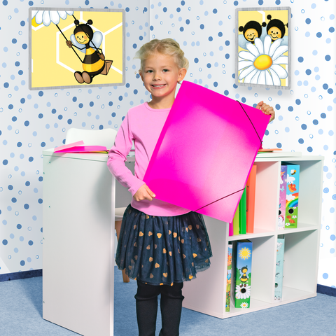 Ein Mädchen mit blonden Locken hält ein großes pinkes Blatt Papier in einem bunten Raum mit Bienenmotiven an den Wänden.