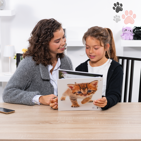 Eine Frau und ein Mädchen sitzen an einem Tisch und betrachten gemeinsam ein Buch mit Bildern von Katzen.