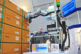 Industrieroboter von Herrmann, der mit Kisten in einem Ausstellungsraum interagiert, im Vordergrund ein Schild mit dem Firmennamen.