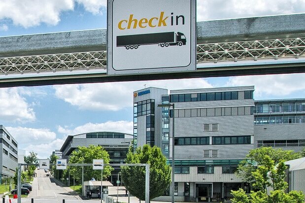 Ein moderner Gebäudekomplex mit einem Überweg, der Verkehrsschilder und ein "Check-in"-Schild zeigt, umgeben von Bäumen und blauem Himmel.