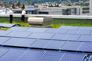 Zwei Personen in Anzügen stehen auf einem Dach mit Solarpanelen, im Hintergrund sind Gebäude und eine Stadtlandschaft sichtbar.