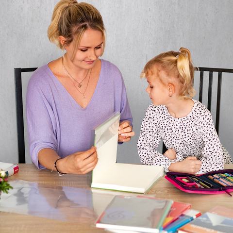 Eine Frau und ein kleines Mädchen sitzen an einem Tisch, während sie gemeinsam an einem Kunstprojekt arbeiten. Bunte Stifte und ein Blumenarrangement sind sichtbar.