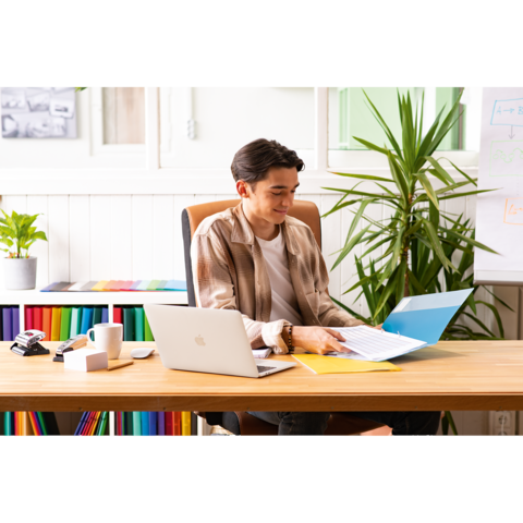 Ein junger Mann sitzt an einem Schreibtisch, arbeitet mit einem Laptop und blättert durch Dokumente in einer Ordner. Pflanzen sind im Hintergrund sichtbar.