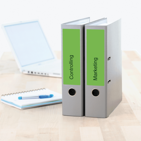 Zwei Aktenordner mit den Beschriftungen "Controlling" und "Marketing" stehen auf einem Tisch, neben einem Laptop und einem Notizblock.