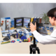 Ein Fotograf steht vor einem Tisch mit verschiedenen Büroartikeln, darunter Ordner, Kleber und Bastelmaterialien, während er eine Kamera hält.