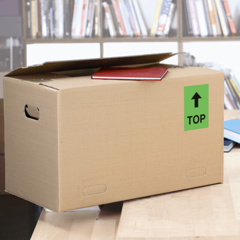 Kartonbox auf einem Tisch, mit einem roten Heft oben drauf und einem grünen Aufkleber mit der Aufschrift "TOP".
