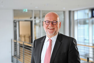 Ein lächelnder Mann mit Brille und Anzug steht in einem modernen Innenraum. Er trägt eine Krawatte in Rosa und hat eine freundliche Ausstrahlung.