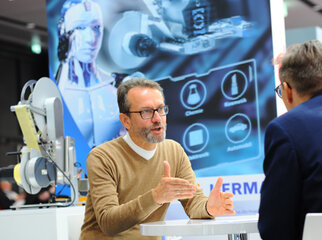 Ein Mann im Gespräch mit einem anderen, während im Hintergrund eine technische Präsentation und ein Roboter sichtbar sind.