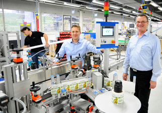 Zwei Männer stehen an einer Produktionslinie in einer Fabrik, während ein dritter Mitarbeiter im Hintergrund arbeitet.