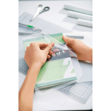 Hände ziehen eine transparente Folie von einem grünen Buch mit der Nummer 2 ab, um es einzuhüllen. Schreibtisch mit Werkzeugen im Hintergrund.