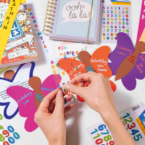 Hände, die bunte Aufkleber auf Schmetterlingsformen anbringen, umgeben von Schulmaterialien und Notizbüchern mit Namen.