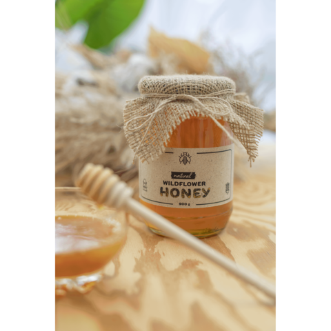 Glas Honig mit Juteverschluss und Etikett "Wildflower Honey" steht neben einem kleinen Glas und einem Honiglöffel auf einem Holztisch.