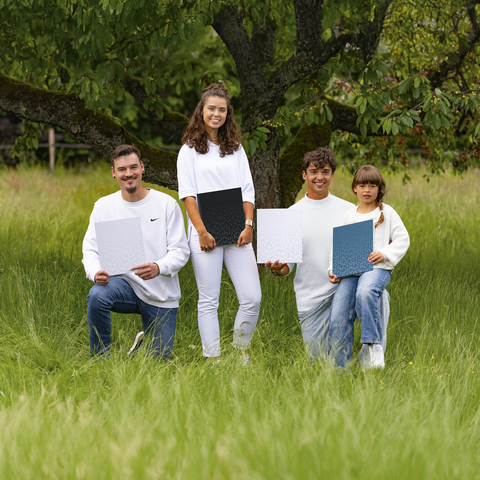 Vier Personen in weißen Oberteilen stehen und sitzen im Gras, halten jeweils Plakate in den Händen. Ein Baum ist im Hintergrund sichtbar.