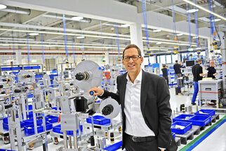 Ein Mann in einem Anzug steht in einer modernen Produktionshalle und hält ein Gerät. Im Hintergrund sind Arbeitsstationen und blaue Behälter sichtbar.
