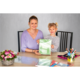 Eine Frau und ein Mädchen sitzen an einem Tisch, halten ein grünes Heft und lächeln. Auf dem Tisch liegen Schreibwaren und Blumen.