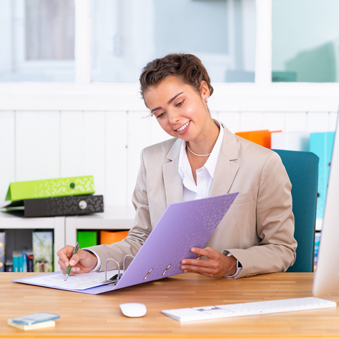 Eine Frau in einem hellen Anzug sitzt an einem Schreibtisch und betrachtet einen Ordner, während sie Notizen macht.