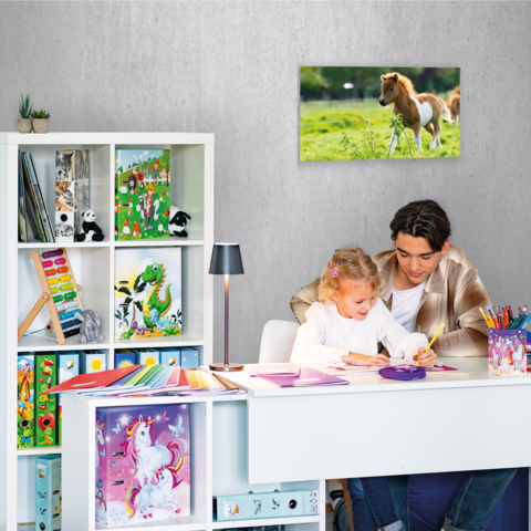 Ein Mädchen sitzt an einem Schreibtisch, malt und schreibt in ein Heft. Im Hintergrund sind Regale mit Spielzeug und Bastelmaterial.