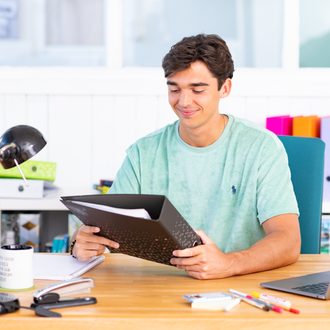 Junger Mann mit lässigem T-Shirt sitzt an einem Schreibtisch und betrachtet einen Ordner, umgeben von Schreibutensilien und einer Lampe.