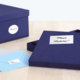 Eine geöffnete, dunkelblaue Geschenkbox mit einem "Happy Birthday!"-Aufkleber und zwei Umschlägen in verschiedenen Farben daneben.