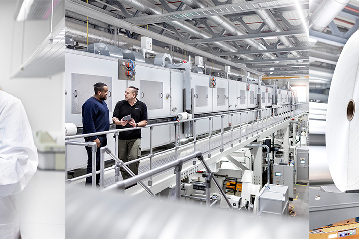 Zwei Frauen in Laborkitteln betrachten ein Dokument, während im Hintergrund eine Produktionslinie mit großen Rollen zu sehen ist.