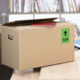 Kartonbox auf einem Tisch, mit einem roten Heft und einem grünen "TOP"-Schild auf der Seite. Hintergrund mit Bücherregalen.