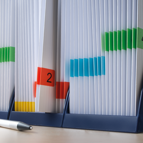 Ordner mit farbigen Markierungen und nummerierten Etiketten, die verschiedene Dokumente organisieren. Ein Stift liegt daneben.