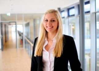 Junge Frau mit langen blonden Haaren, die in einem modernen Bürogebäude in einem schwarzen Blazer und einem weißen Hemd lächelt.