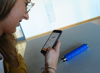 Eine Person mit Brille schaut auf ihr Smartphone, während ein blauer Stift auf dem Tisch liegt.