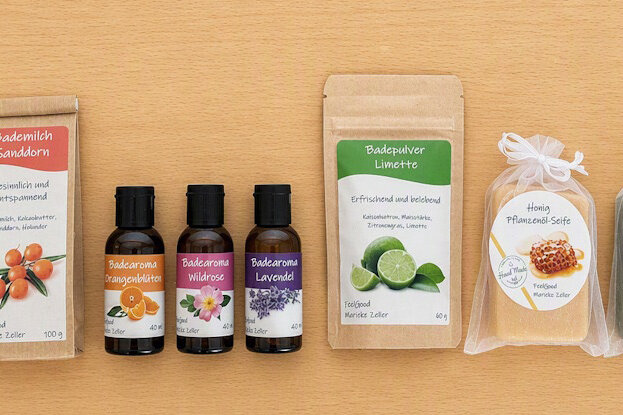 Individuelle Etiketten für Kosmetikprodukte in einer Reihe: Pflanzenöl-Seifen in Klarsichtbeuteln, Doypacks mit Bademilch und Badepulver sowie Braunglasflaschen mit Badearoma, auf einer hellen Holzoberfläche.