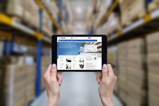 Tablet in Händen, zeigt eine Webseite mit Informationen über Lagerverwaltung, im Hintergrund Regale mit Kartons.