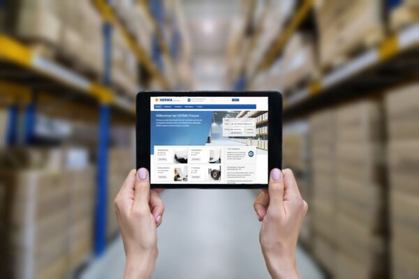 Tablet in Händen, zeigt eine Webseite mit Informationen über Lagerverwaltung, im Hintergrund Regale mit Kartons.