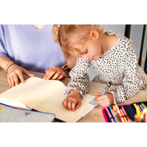 Ein kleines Mädchen schreibt in ein Notizbuch, während eine erwachsene Person daneben sitzt und beobachtet. Bunte Stifte liegen auf dem Tisch.