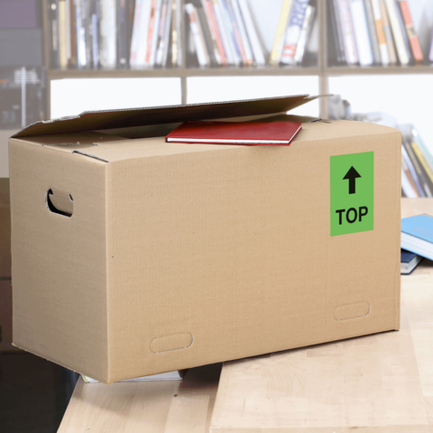 Kartonbox mit geöffnetem Deckel, auf einem Tisch platziert, mit einem roten Objekt und einem grünen Aufkleber, der "TOP" anzeigt.