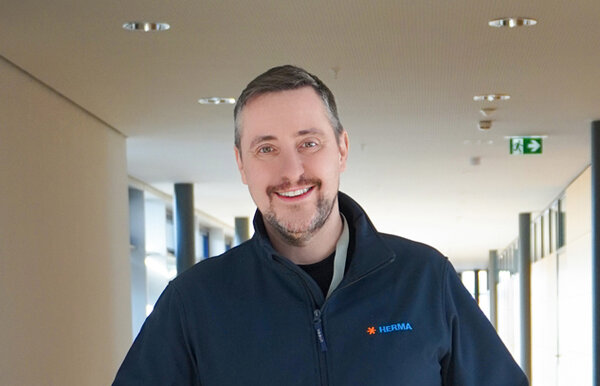 Ein Mann mit kurzem Haar und einem Bart trägt eine schwarze Jacke mit dem Logo von HERMA. Er steht in einem modernen Flur.