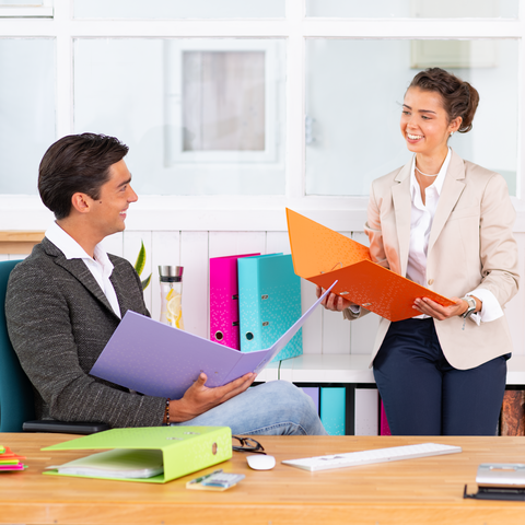 Zwei Personen in einem Büro, die sich unterhalten. Eine Person hält orangefarbene Ordner, während die andere einen lila Ordner in der Hand hat.
