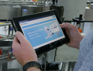 Eine Person hält ein Tablet, auf dem die Webseite des HERMA Service-Portals mit einem Willkommensgruß und einem Roboter-Design angezeigt wird.