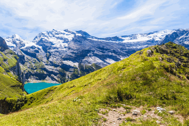 Panoramablick auf grüne Hügel und schneebedeckte Berge, umgeben von klarem Wasser und blauem Himmel.