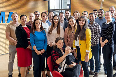 Eine Gruppe von Mitarbeitern steht vor einer Wand mit dem Schriftzug "HERMA". Sie lächeln und posieren gemeinsam in einem modernen Büro.