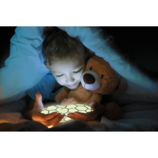 Ein Kind sitzt im Bett, hält ein leuchtendes Objekt in den Händen und umarmt einen Teddy, während es unter einer Decke liegt.