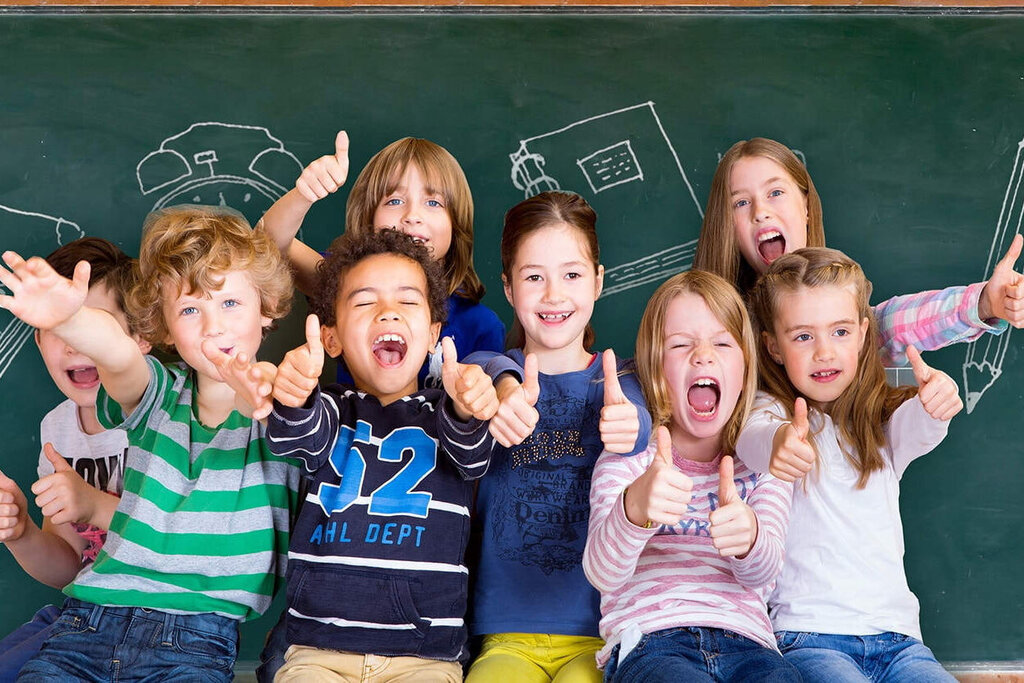 Eine Gruppe von Kindern steht vor einer Tafel, lächelt und posiert fröhlich. Zeichnungen von Schulmaterialien sind im Hintergrund sichtbar.
