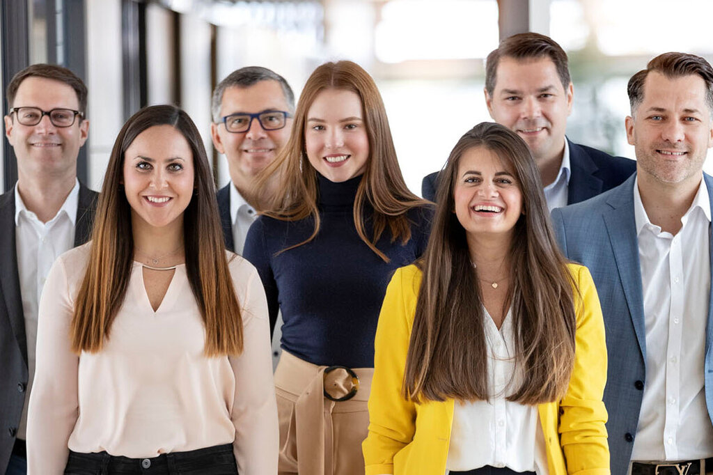 Eine Gruppe von sieben Personen steht in einem modernen Flur. Sie tragen formelle Kleidung und lächeln in die Kamera.