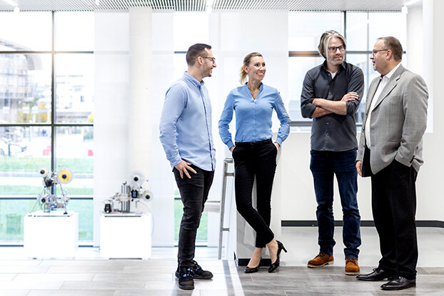 Vier Personen in einem modernen Büro sprechen miteinander, während im Hintergrund ein industrielles Gebäude und eine weitläufige Anlage sichtbar sind.