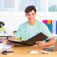 Ein junger Mann sitzt an einem Tisch und betrachtet ein Dokument. Er trägt ein grünes T-Shirt und lächelt, während er Notizen macht.