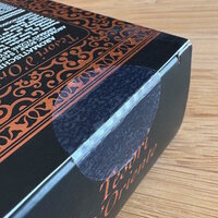 Lösungen Kosmetik: Schwarze Verpackung mit dekorativen, orangen Mustern und Text. Die Lasche der Verpackung ist mit einem transparenten Verschlussetikett versiegelt