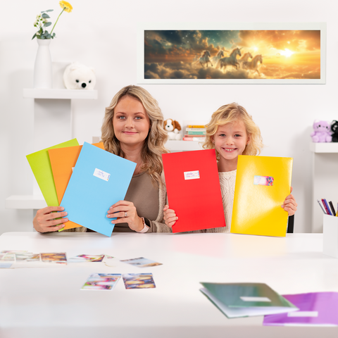 Zwei Mädchen sitzen an einem Tisch und halten farbige Ordner. Auf dem Tisch liegen Fotos und im Hintergrund ist ein Bild von Einhörnern.