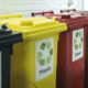 Drei Abfallbehälter in verschiedenen Farben: Gelb für Plastik, Rot für Glas und Schwarz für Restmüll, mit Recycling-Symbolen.