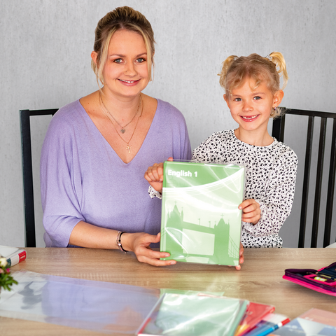 Eine Frau und ein Mädchen sitzen an einem Tisch und halten ein grünes Heft. Auf dem Tisch liegen bunte Stifte und ein Blumenstrauß.