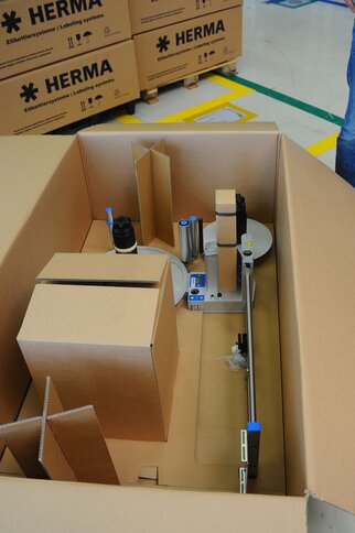 Ein Karton mit einem Etikettiergerät und verschiedenen Kartonagen, das für Verpackungszwecke vorbereitet ist.