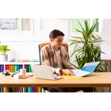 Junger Mann sitzt an einem Schreibtisch, arbeitet mit einem Laptop und blättert durch Dokumente in einer bunten Büroumgebung.